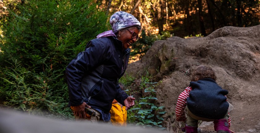 A senior woman in active outdoor clothing encourages a toddler to navigate uneven ground in a lush forest environment.