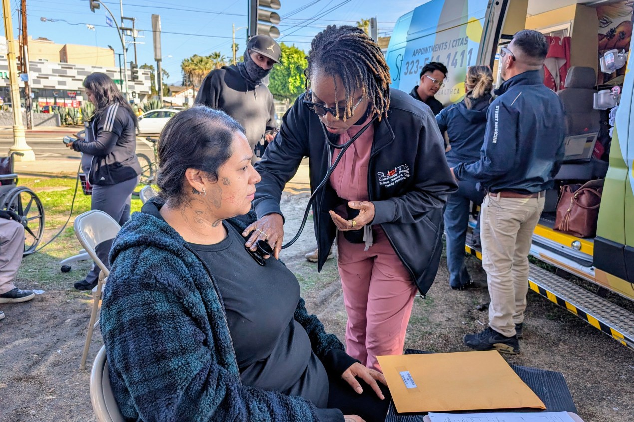 A street medicine nurse holds the end of a stethoscope to a woman's chest.