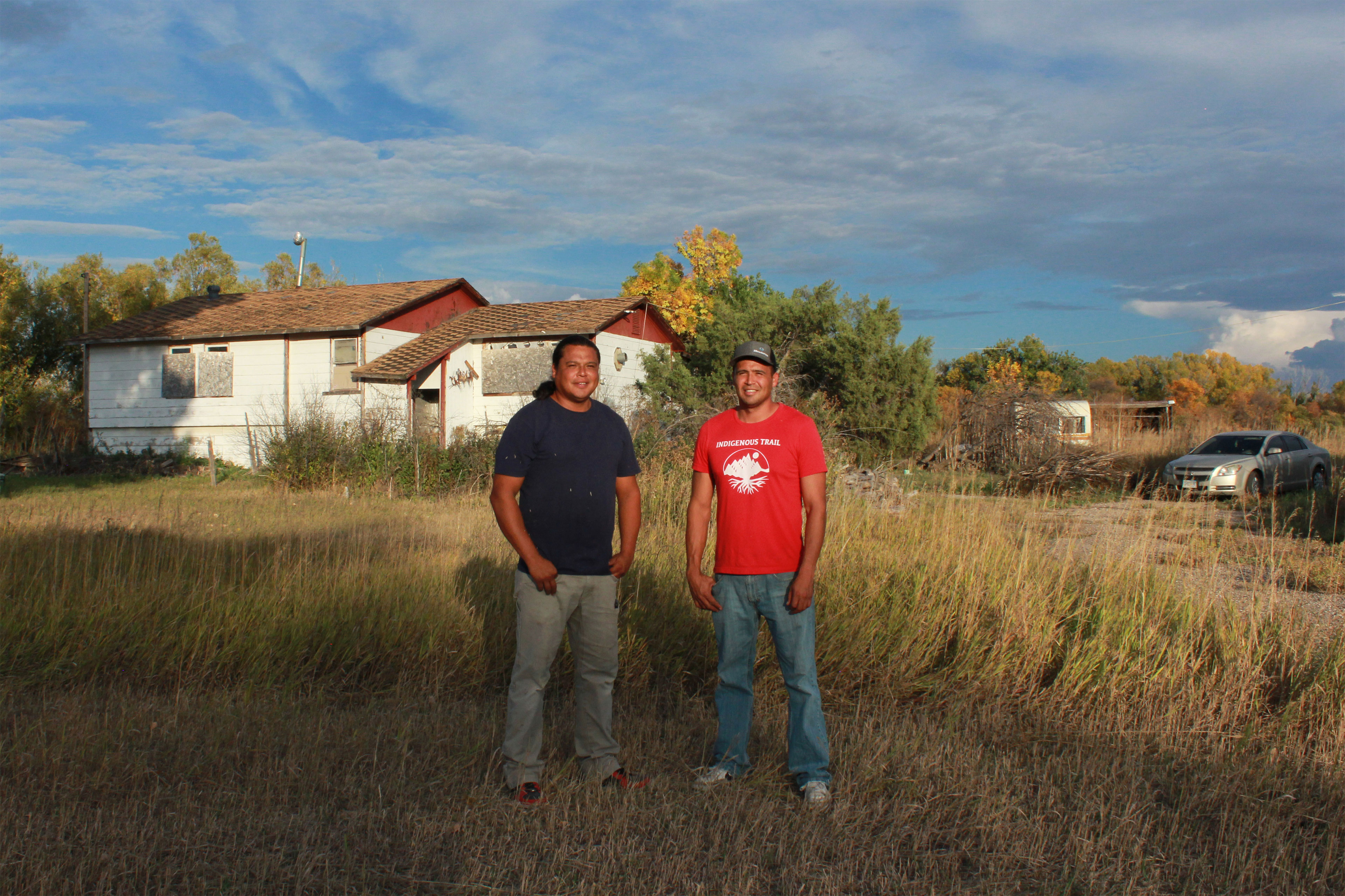 Two men in t-shirts and jeans stand in the grass near a boarded-up house and smile at the camera.