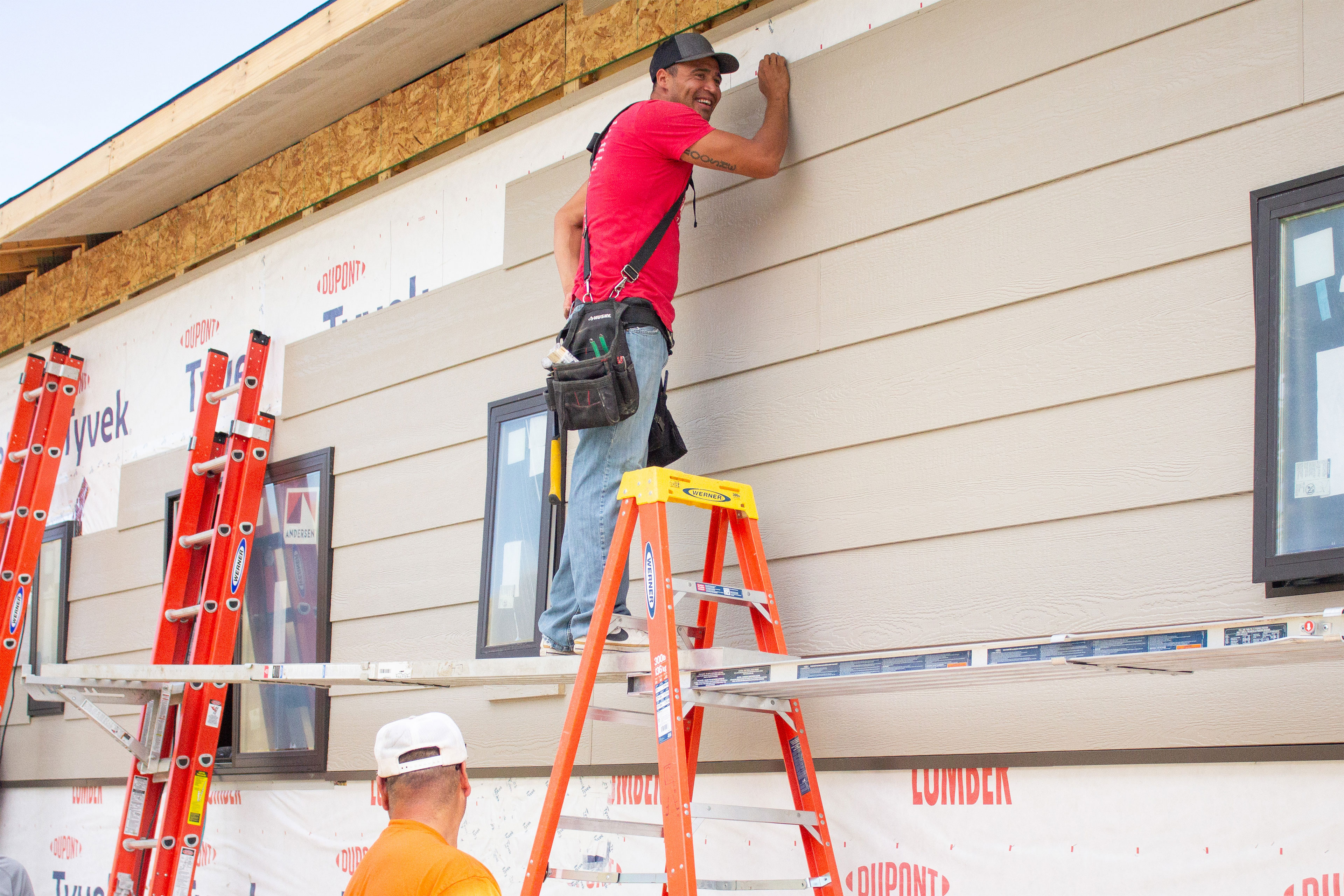 A man wearing a baseball cap, red t-shirt, jeans, and a bag with tools in it, stands on makeshift scaffolding between ladders and holds a panel to the side of a building under construction.