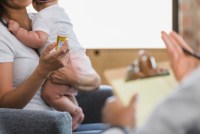 An unrecognizable new mom holds her baby in one arm, and a pill bottle in the other. She is talking with a medical professional.