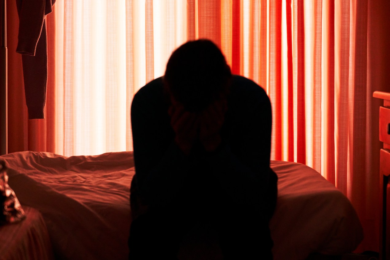 A person sitting on a bed, their head in their hands, backlit by a window