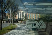 A building seen through the glass of a wind shield next to an outdoor bench. An etching in the glass reads "FDA" and the sign above the entrance to the building in the distance reads "Food and Drug Administration."