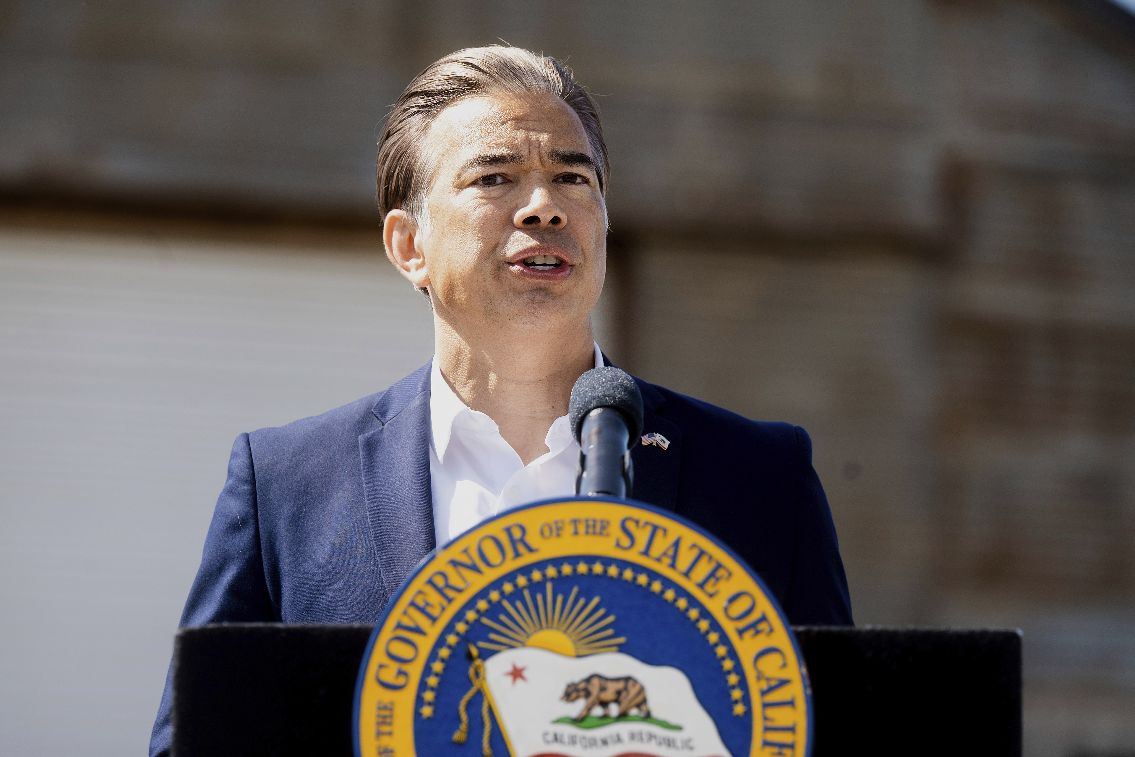 A man in a navy blazer and white collared shirt stands at a podium with a microphone