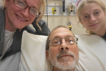 An older man lies in a hospital bed. His wife and daughter are on either side of him, smiling.